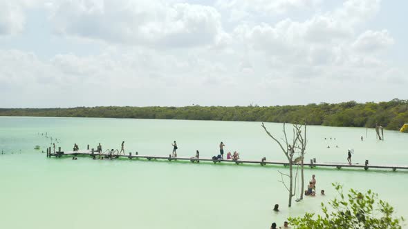 Fly Over People Relaxing at Water Sitting at Long Wooden Pier or Bathing in Natural Swimming Pool
