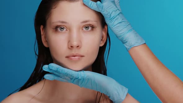 Beauty Surgeon Arms in Blue Gloves Touching Woman Face Isolated on Blue