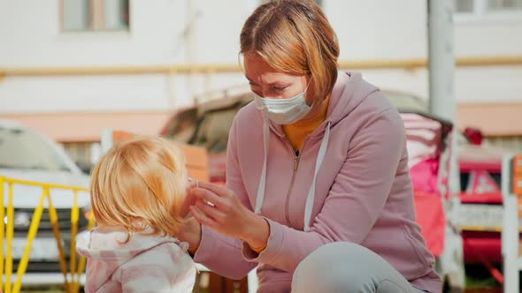 A mother puts a medical mask on her little child sitting on a children's slide.