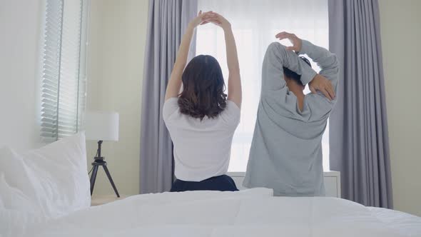 Rear view of Asian couple stretching hands after wakeup from bed in bedroom together in morning.