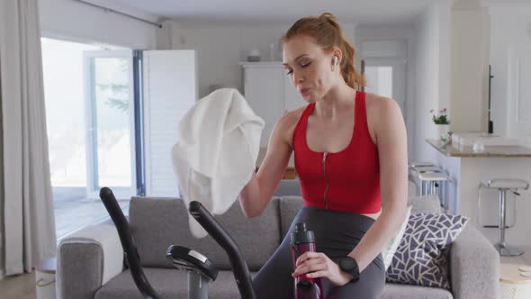 Woman drinking water and wiping her face with a towel while sitting on stationary bike at home