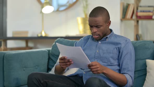 Young African Man with Documents Reacting to Loss on Sofa