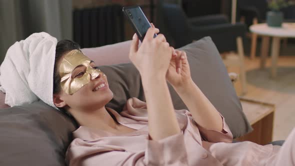 Pretty Woman In Gold Sheet Mask With Smartphone