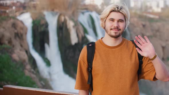 A Young Blonde Smiling Man Stands on the Background of Waterfall and Waving His Hand