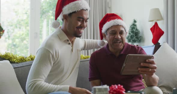 Cheerful biracial son with senior father in santa hats making video call on tablet pc in christmas