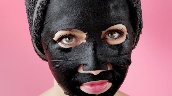 Young woman apply black cosmetic fabric facial mask on pink background. Face peeling mask with charc