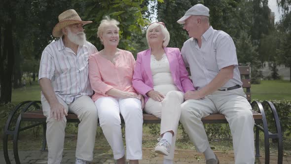 Two Cute Mature Couples Talking and Smiling Sitting on the Bench in the Summer Park