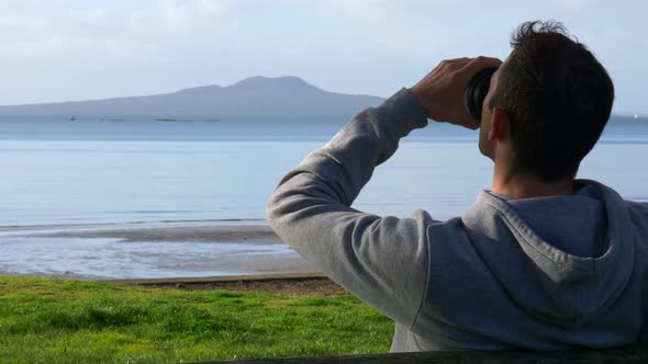 Young caucasian man drinking coffee sitting on a bench at the beach  looking at Rangitoto island in