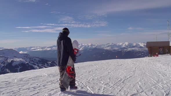 A young man snowboarder walking with his board on a scenic snow covered mountain top.
