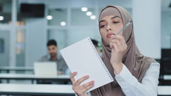 Pensive Young Arabic Female Worker in Hijab Sitting at Office Desk Using Paper Notebook Making Notes