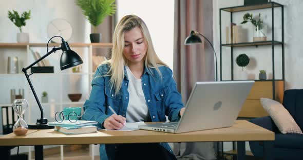 Blond Young Woman Working with Laptop and Doing Needed Notes Into Papers in Home Office