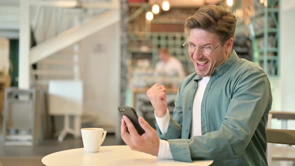 Middle Aged Man Celebrating Success on Smartphone in Cafe 