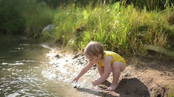 Little Funny Cute Blonde Girl Child Toddler in Yellow Wet Bodysuit Playing By the Lake Waterside