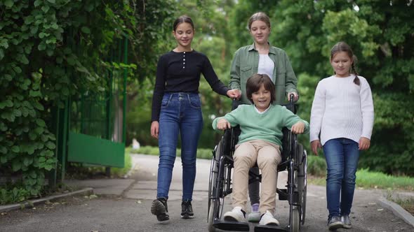 Front View Cheerful Girls Pushing Wheelchair with Happy Caucasian Disabled Boy Talking Walking in