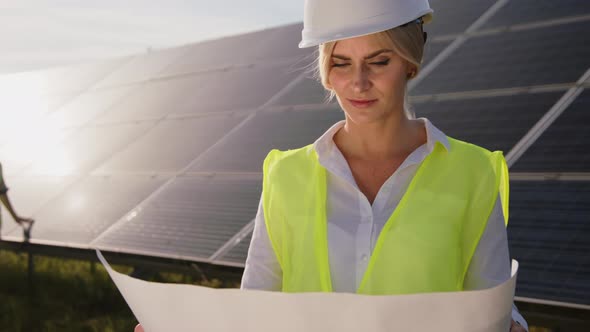 Beautiful Female Solar Engineer Standing Near the Panels with a Drawing on Paper and a Young