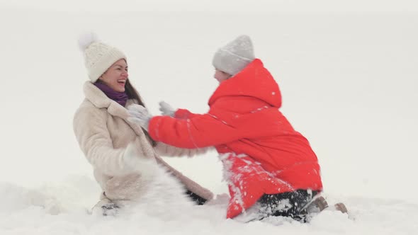 Young Woman with Her Teenage Daughter is Kneeling in the Snow and Cheerfully Sprinkling Snow on Each