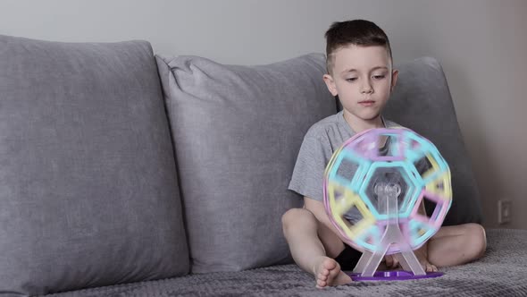 A boy in a gray T-shirt sits on a bed near a white wall and plays with a Ferris wheel alt