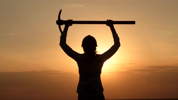 Silhouette woman in helmet stands outdoors background sunset and raises pickaxe over head, back view