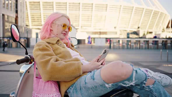 Young Stylish Woman with Pink Hair Relaxing on Scooter and Web Surfing Social Media on Smartphone