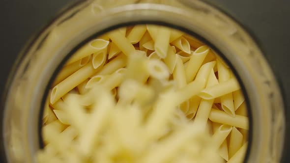 TOP VIEW: Uncooked Penne Rigate Fall Into Glass Jar On Black Background - Slow Motion