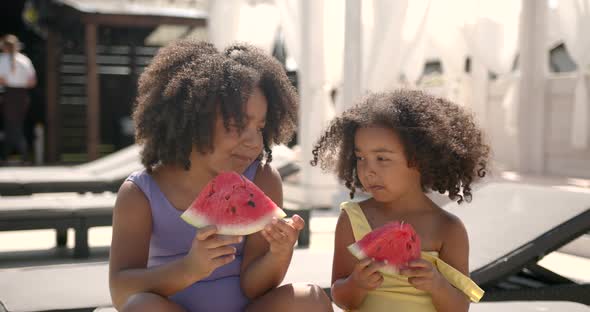 Two Cute Girls Eating Watermelon on Loungers at Pool