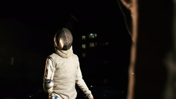 Two Fencers In Fencing Suits And Helmets Fencing In The Dark Outside.