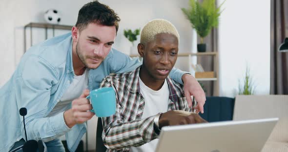 Smiling Young Mixed Race Gays Talking with Each other while Applying Laptop at home