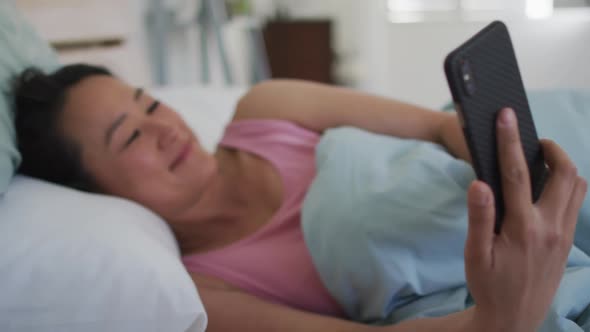 Happy asian woman lying in bed, using smartphone in the morning