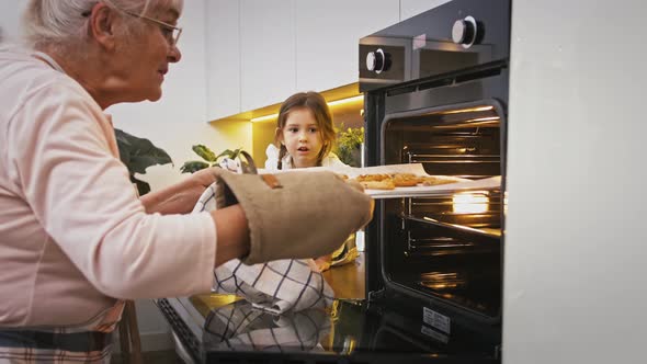 Grandmother is Taking Out Hot Cookies From Oven Surprised Granddaughter is Looking at It