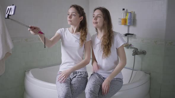Charming Twin Sisters Sitting on Bathtub Taking Selfie on Smartphone Smiling