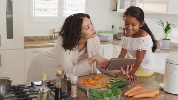 Hispanic mother teaching daughter sitting on countertop cooking, looking at tablet