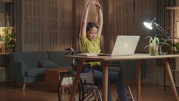 Asian Kid Girl Sitting In A Wheelchair Stretching While Using Laptop Computer At Home