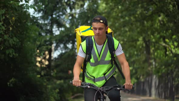 Portrait of Young Caucasian Delivery Man in Green Vest with Yellow Backpack Riding Bicycle Gesturing