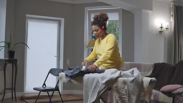 Wide Shot of Joyful African American Woman Ironing Laundry Dancing and Singing to Music in