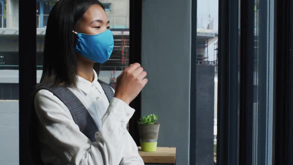 Thoughtful asian woman wearing face mask looking out of window at modern office