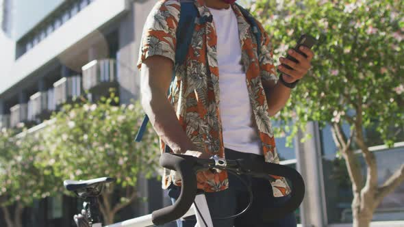 Midsection of african american man in city, wheeling bike and using smartphone in the street