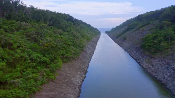 Aerial backwards shot of small river dam surrounded by rocky shore and green plants growing on hill