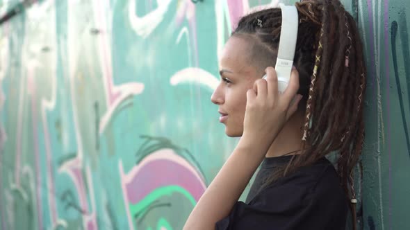 Young Modern Woman Listens to Music on Headphones Outdoors Background of a Wall with Graffiti