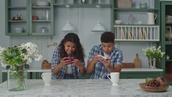 Young Adult African American Couple Playing Mobile Game on Mobile Phones Sitting in Kitchen