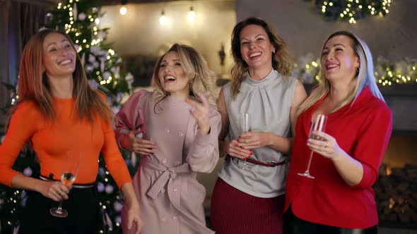 Four Girlfriends in Elegant Clothes Dance with Glasses in Hands in a Room Decorated for Christmas