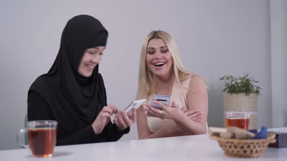 Happy Caucasian and Muslim Women Using Smartphones and Laughing. Cheerful Female Friend From