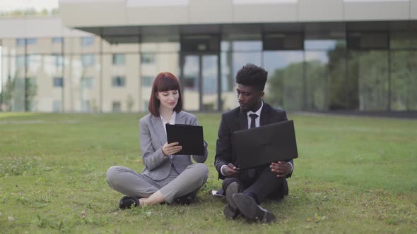 Two Business Colleagues in Formal Clothes Sitting on Grass Outdoors and Signing the Contract