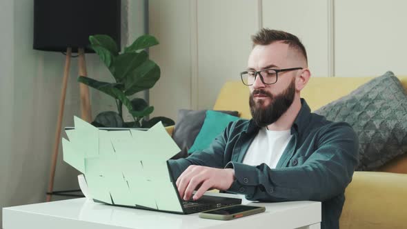 Man Working at the Computer with a Lot of Reminder Notes Sitting Near Sofa