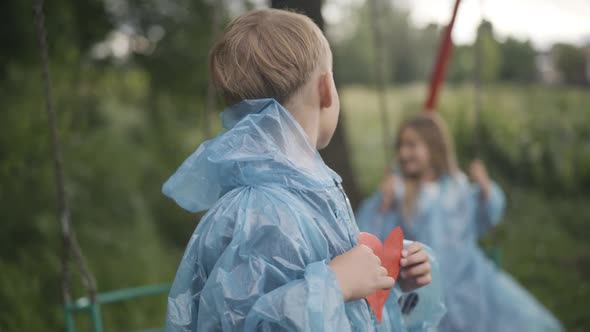 Confident Caucasian Boy with Paper Heart Looking Back at Girl Swinging on Swings and Smiling at