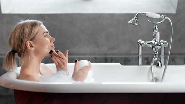 Smiling Young Natural Girl Taking Spa Foam Bath Applying Face Cream Side View