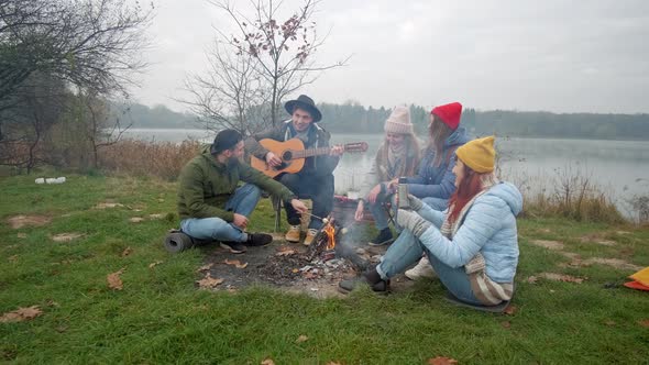 Happy Friends Sitting Around a Bonfire on the Beach, Playing Guitar and Singing