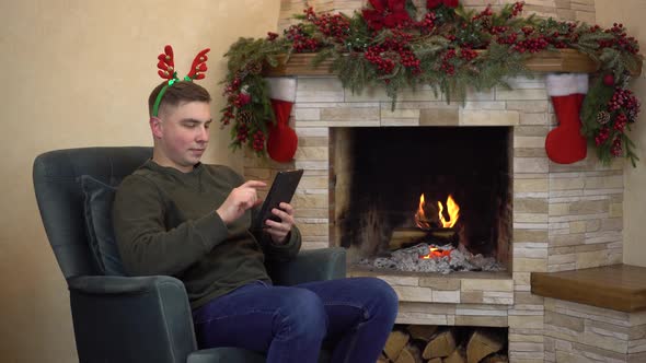 A Young Man is Sitting in a Chair with Horns on His Head By the Fireplace and Texting From a Tablet