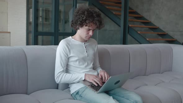 Concentrated Curly Guy Typing on Laptop While Sitting on Sofa in Fashionable Apartment