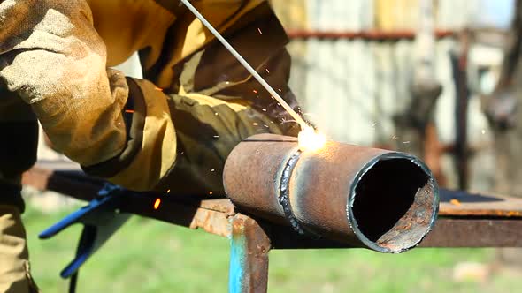 Male Welder in Protective Suit and Mask is Welding a Thick Steel Pipe with an Electrode on an Iron
