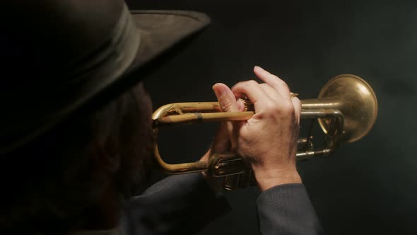 Musician in Hat and Jacket Plays Copper Pipe on Dark Background in Light of Searchlight Top View